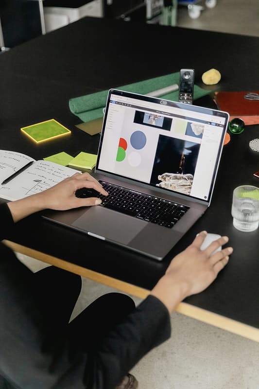 Someone works on a laptop displaying colorful design elements amid creative tools and notes